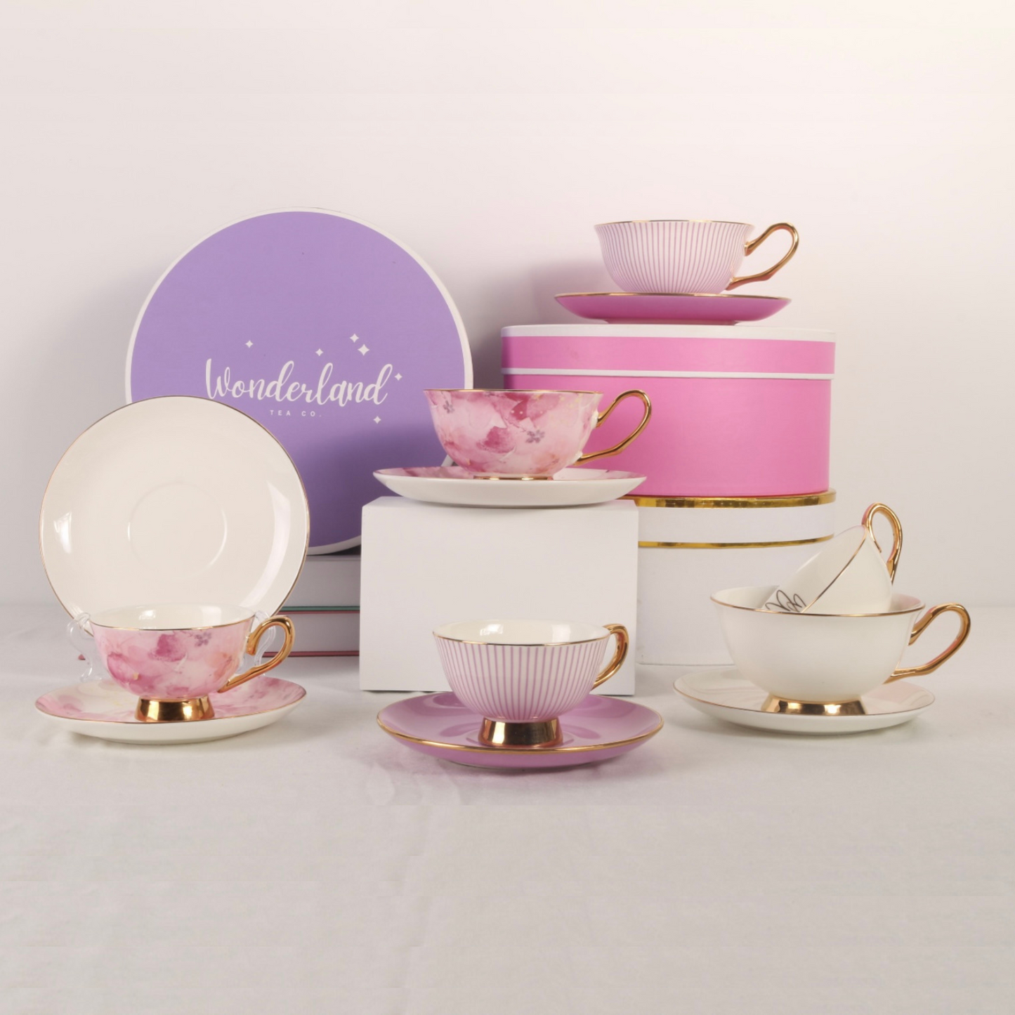 Six teacups arranged on table with giftboxes. Small and standard teacups with matching saucers. Colours include pink flowers, purple stripes and white with gold hearts. All teacups have gold handles and edges.