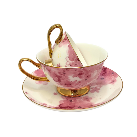 Two teacups with pink flowers on a white background with gold handle and edges. Mini teacup stacked inside standard sized teacup.  Sitting on matching pink flower saucer.  Isolated on a white background. 