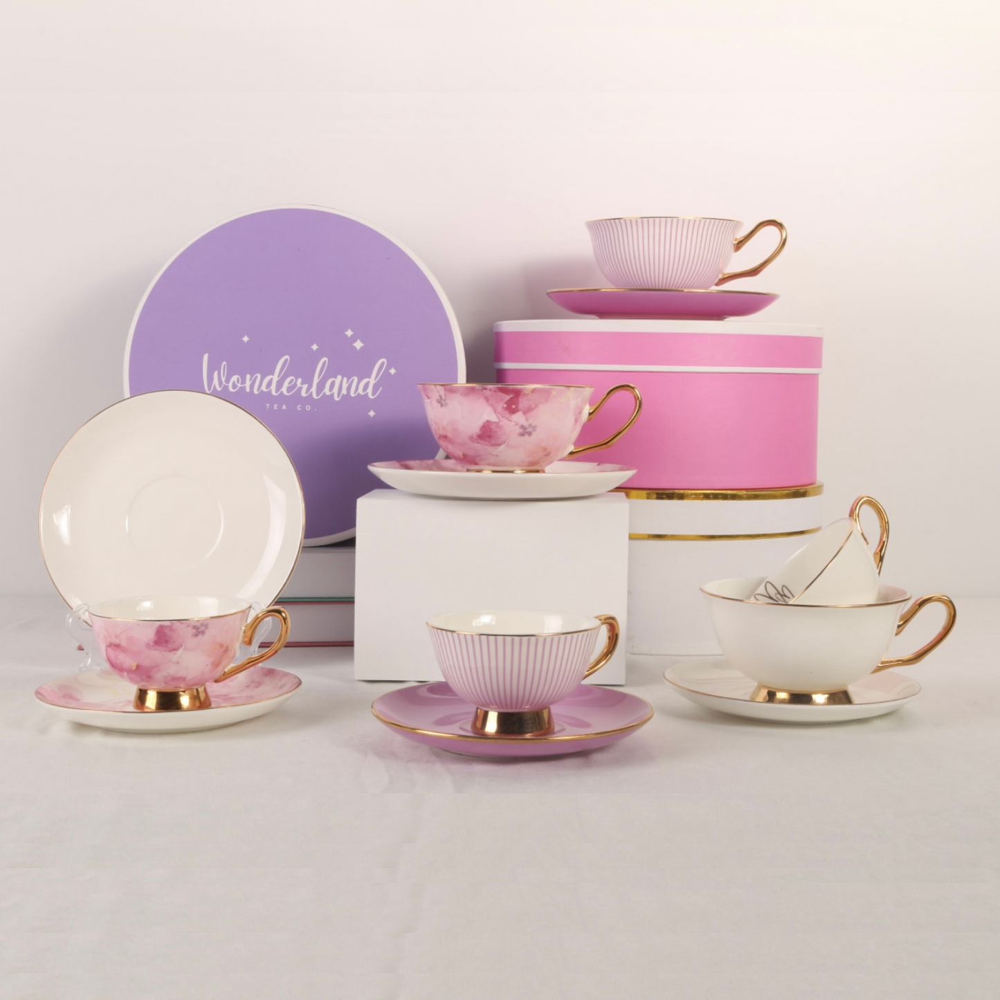 Six teacups arranged on table with giftboxes.  Small and standard teacups with matching saucers.  Colours include pink flowers, purple stripes and white with gold hearts.  All teacups have gold handles and edges.